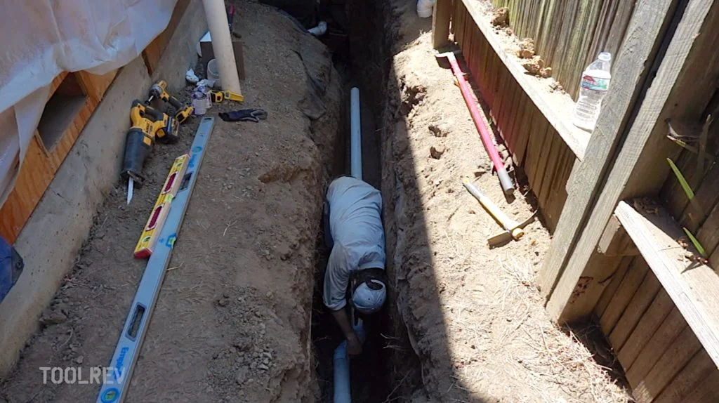 Worker in a trench laying white PVC pipe beside a fence