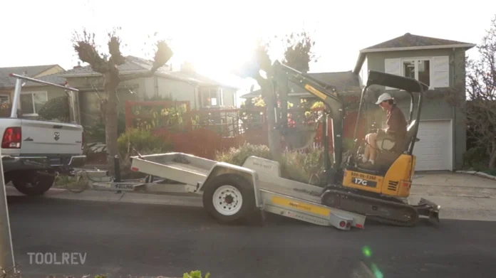 Rental mini excavator on a trailer behind a pickup truck in a driveway